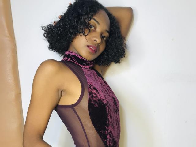 HotPoupee69 com cabelo preto encaracolado, vestindo um vestido de alça roxo de veludo, braços levantados, olhando para a câmera, mostrando os seios através de um tecido transparente, batom vermelho, fundo branco..