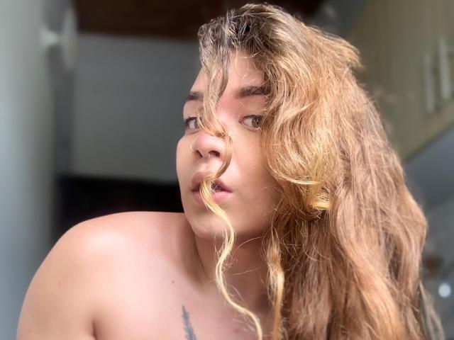 BlueSofy with wavy blonde hair, looking slightly to the side, showing her bare shoulders and a small tattoo on her chest. Her face has a subtle, pouty expression.