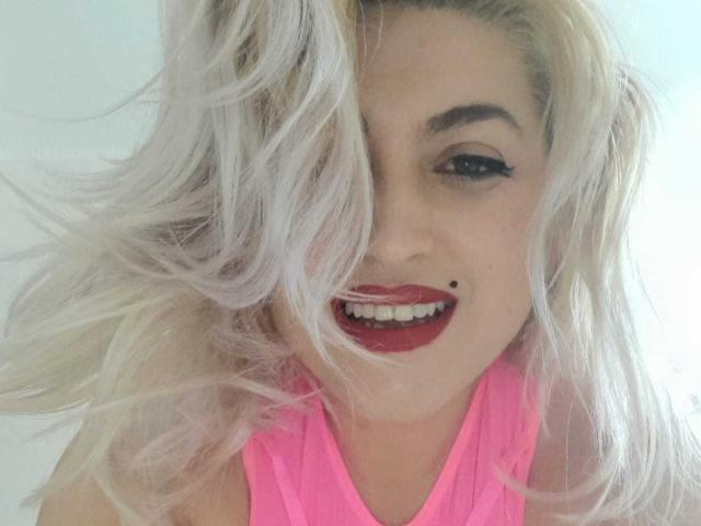 Elizabeth Raegan, met blond platinakleurig haar, rode lippenstift en een zwarte moedervlek boven haar licht geopende mond, draagt een roze top en glimlacht naar de camera..