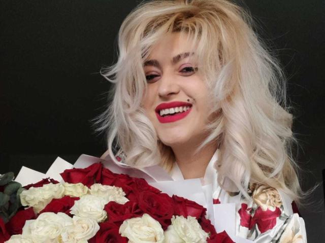 Elizabeth Raegan, con cabello rubio y ondulado, lápiz labial rojo, sonriendo, sosteniendo un ramo de rosas rojas y blancas, vestida con una camisa blanca con estampados florales, luciendo adorable..