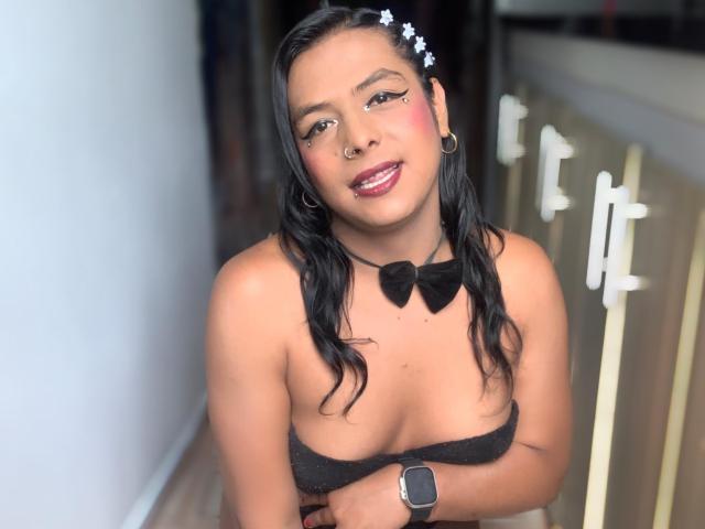 EmilyTsSweet with black hair, star headband, nose ring, earrings, bow tie, and black top, covering her tits, smiling, wearing a watch, in a hallway.