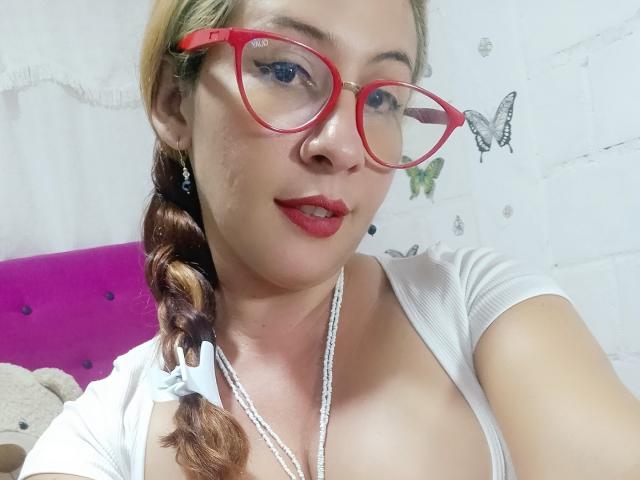 NatyBoobs con gafas rojas, camisa blanca y collar, mostrando sus senos, labios rojos, cabello trenzado y ojos azules, sonriendo ligeramente. Pared beige con calcomanías de mariposas..