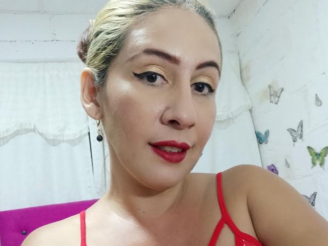 NatyBoobs con capelli biondi, rossetto rosso e eyeliner nero, indossa un vestito con spalline rosse, guarda la fotocamera con un leggero sorriso, in una stanza bianca con farfalle..