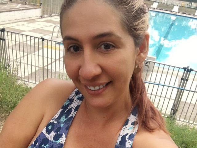 NatyBoobs, une femme souriante aux cheveux bruns attachés en queue de cheval, portant un haut à enfiler bleu et blanc à motifs, montrant ses seins, se tient près d'une piscine entourée d'une clôture..