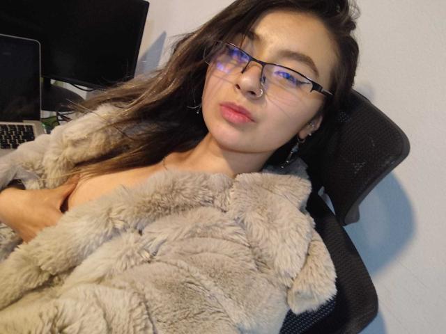 EymyFox with glasses, pouty lips, and brown hair, wearing a fluffy gray robe, sitting in a chair, holding her tits, looking at the camera, in a relaxed pose.