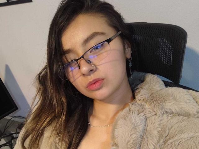 EymyFox with glasses, pouty lips, brown wavy hair, fur-lined jacket, ear piercings, sitting in black chair, reflection on glasses.
