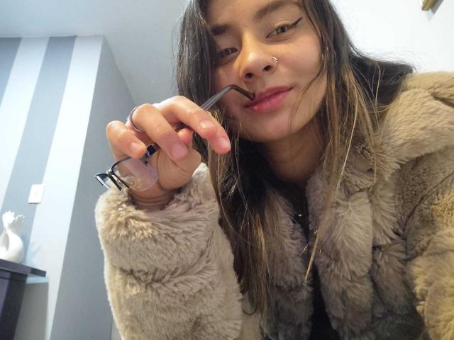 EymyFox with long brown hair, wearing a furry beige coat, applying black eyeliner with a precise line, has a nose ring, and a subtle smile.