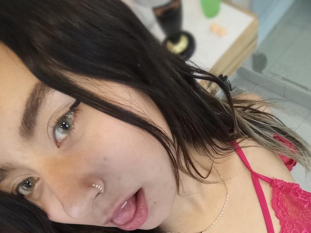 EymyFox has black hair, a nose ring, and a septum piercing. She's sticking out her pink tongue, wearing a red lace top, and has green eyes.
