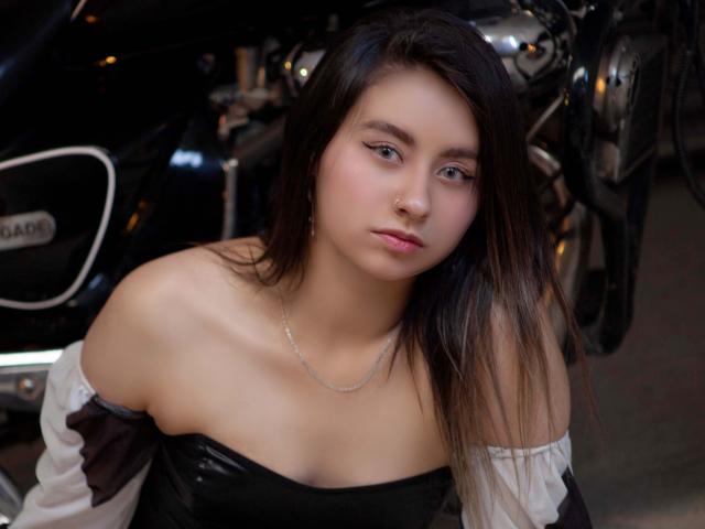 EymyFox has pale skin, long brown hair, and small tits, wearing a black off-shoulder top with white sleeves, standing in front of a shiny black motorcycle.