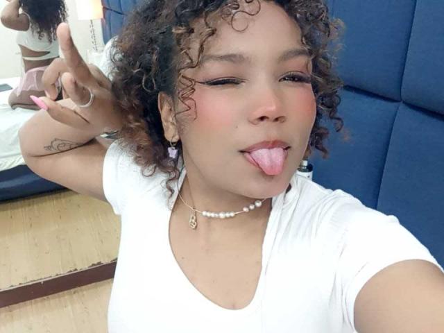 EvaBennet has curly brown hair, light brown skin, and a white shirt. She's sticking her tongue out, flashing a peace sign, with a pearl necklace and ring.