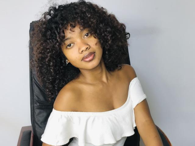 CandyMagicX with curly dark hair, brown eyes, and light brown skin, wearing an off-shoulder white top, sitting in a black chair, looking slightly to the left.