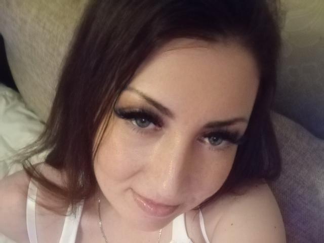 WikiStar has fair skin, brown hair, and blue eyes with long, thick, black eyelashes. She's smiling slightly with a subtle sheen on her face, wearing a white top.