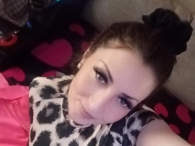 WikiStar with dark hair in a black bun, wearing a black and white patterned top, pink shirt, and heart-patterned black blanket, looking up with pouty lips.