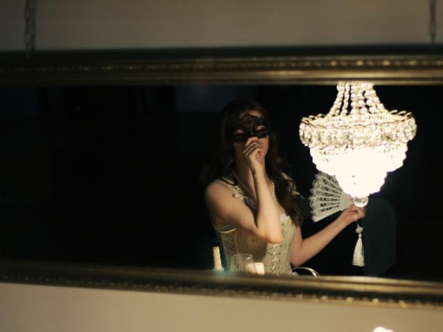 SwetMila with dark brown wavy hair, wearing a black lace mask, and a beige corset, covering her mouth in a reflective gold-framed mirror with a crystal chandelier.