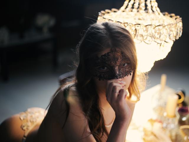 SwetMila with brown hair, wearing a black lace mask, looking seductively at the camera, resting her chin on her hand, under a glowing crystal chandelier. Her ass is visible.