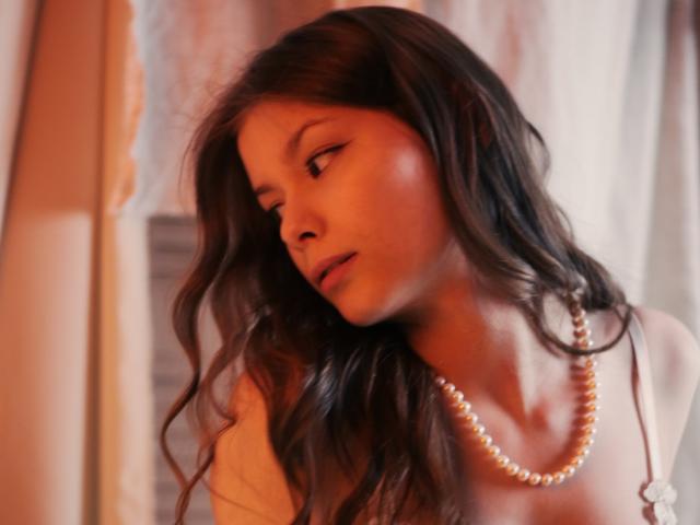 SwetMila with long brown wavy hair, wearing a pearl necklace, looking down with a pensive expression, soft orange lighting on her face, blurred background, upper body shot.