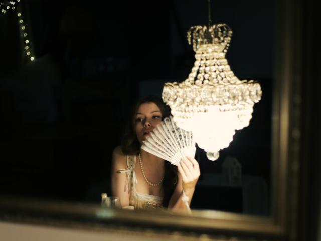 SwetMila with tits showing, holding a white fan, wearing a pearl necklace, under a glowing crystal chandelier, looking seductive, in a dark room, reflected in a mirror.