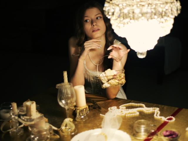 SwetMila with tits up, holding a fork, wearing a pearl necklace, looking at a chandelier, table with candles, glass, and jewelry in a dimly lit room.
