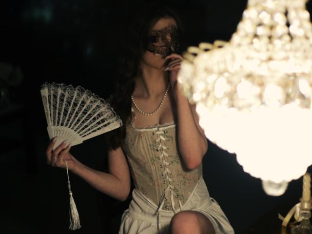 SwetMila with dark curly hair, wearing a lace mask, beige corset, pearl necklace, holding a white fan, sitting near a glowing crystal chandelier. Her tits are visible under the corset.