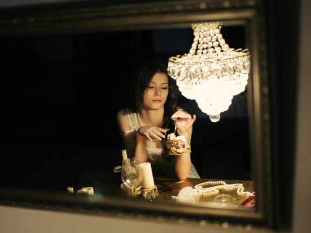 SwetMila with tits, wearing a pearl necklace, sits under a chandelier, eating from a plate in a mirror's reflection, with candles and jewelry on a wooden table.