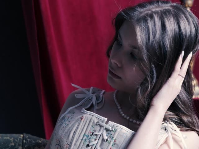 SwetMila with wavy brown hair, wearing a white floral dress and pearl necklace, touches her head with a soft expression, red curtain in the background.