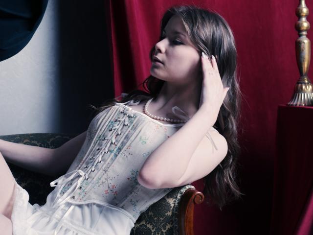 SwetMila with pale skin, brown hair, wearing a white floral corset, touching her head, sitting on an ornate green chair, red curtain, gold lamp, looking to the side.