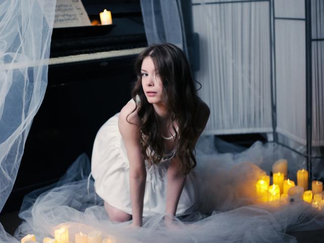 SwetMila with long brown hair, white dress, and pearl necklace, kneeling on a gray floor with lit candles, surrounded by white tulle, looking to the side with a serious expression.