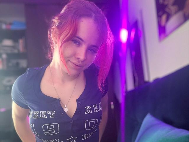 NiaSpencer, com cabelo rosa, sorrindo, vestindo uma camisa azul escura com a inscrição "Neet", mostrando seus pequenos seios, e um colar com um pingente de coração. Luz LED roxa ao fundo..