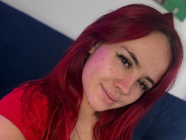 Nia Spencer hat rotes Haar, ein süßes Lächeln und Sommersprossen, trägt ein rotes Hemd und hat eine leichte, violette Prellung auf ihrer rechten Wange. Sie schaut in die Kamera..