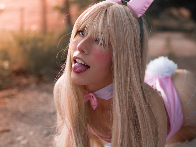 ValerieBunny with blonde hair, pink bunny ears, tongue out, pink blush, and a pink bow, wearing a pink bunny suit, on all fours, in soft outdoor light.