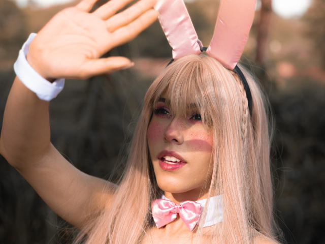 ValerieBunny with pink bunny ears, white cuffs, pink bowtie, and long blonde hair, waving with hands up, showing pink makeup and smiling with glossy lips.