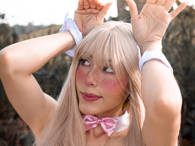 ValerieBunny with blonde hair, pink bowtie, white cuffs, hands up, looking sideways, pink blush, freckles, smiling, pink lipstick, bare shoulders, rock background, sunlight.
