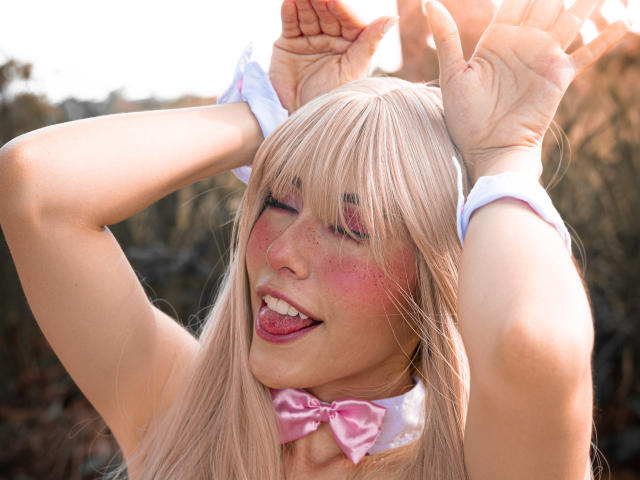  ValerieBunny with blonde hair, pink bowtie, and white wristbands, sticking out tongue, hands raised, eyes closed, blushing, freckles, sunlight, outdoors, playful expression, soft pink makeup.