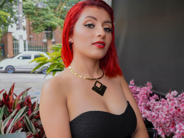 LizBonnet with bright red hair, black strapless top, gold necklace with black geometric pendant, red lipstick, and black earrings, standing near pink flowers and green plants.