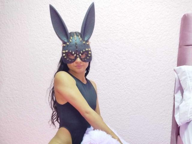 CrisyMarie with black mask, large black bunny ears, and black one-piece swimsuit, holding fluffy white toy, sitting in pink room, pink headboard, long black hair.