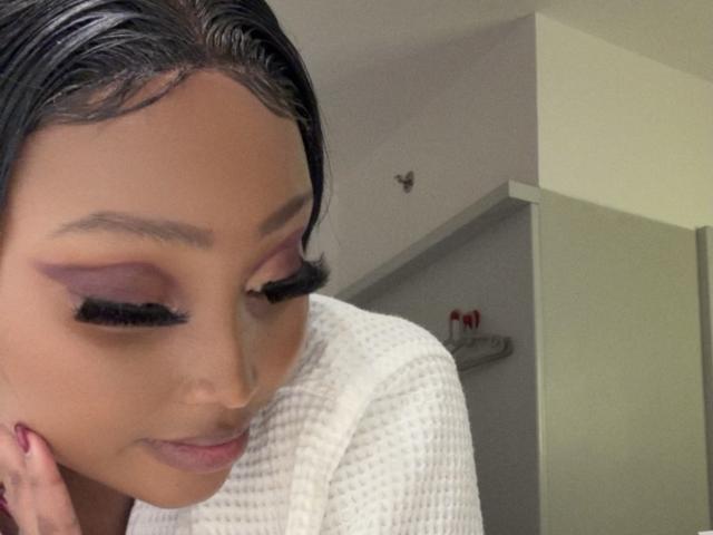 CassieYk with dark hair, brown skin, and purple eyeshadow, wearing a white waffle-texture top, has long black eyelashes and a subtle smile, resting her chin on her hand.