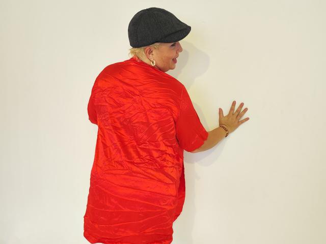RebeccaSinss, con cabello rubio, usando un gorro negro plano y una camisa roja brillante y arrugada, está de pie con las manos apoyadas contra una pared blanca, mirando de lado. Tiene un trasero regordete..