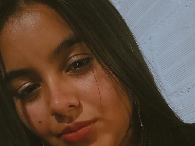 AmberMedellin's face with dark, straight hair, brown eyes, nose ring, and a slight smile, showing a few moles and freckles, looking down with a soft, relaxed expression.