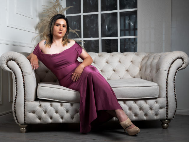 LiliEvanss with medium tits, wearing an off-shoulder purple dress, sitting on a tufted beige sofa, legs crossed, beige heels, relaxed, blonde hair, subtle makeup, confident expression.