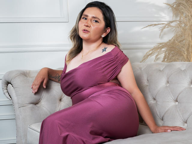 LiliEvanss with medium tits, sitting on a beige couch, wearing a purple off-shoulder dress, black star tattoo on left shoulder, light skin, wavy blonde hair, pearl earrings.