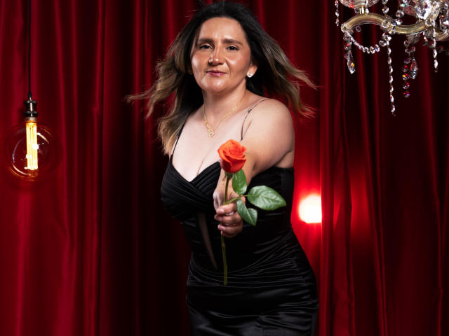LiliEvanss with medium tits, black satin dress, holding a red rose, smiling, wavy brown hair, red curtain, hanging light bulb, and a crystal chandelier in the background.