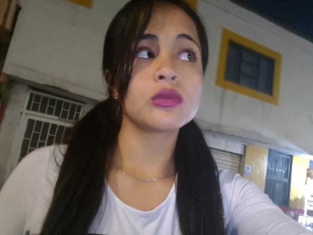 EmilyBennitt has dark hair in pigtails, pink lipstick, and a nose ring, looking up with a pouty expression, wearing a white shirt, in a selfie.