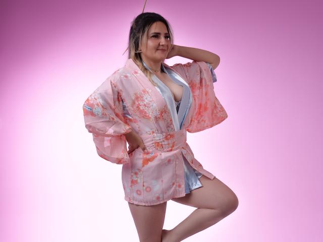 LiliEvanss wearing a pink floral kimono with a blue inner lining, exposing her tits, with one leg raised, hand on her head, smiling, against a pink gradient background.