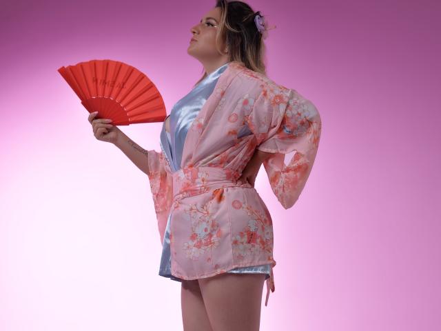 LiliEvanss with pink floral kimono, red fan, and blue silk top, holding fan in right hand, looking up, pink background, blonde hair, flower in hair, sexy, confident.