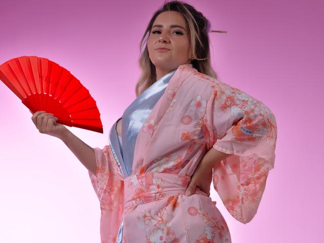 LiliEvanss in a pink floral kimono, holding a red folding fan, with a hairpin in her blonde hair, looking confident. Her tits are partially visible under the open kimono.