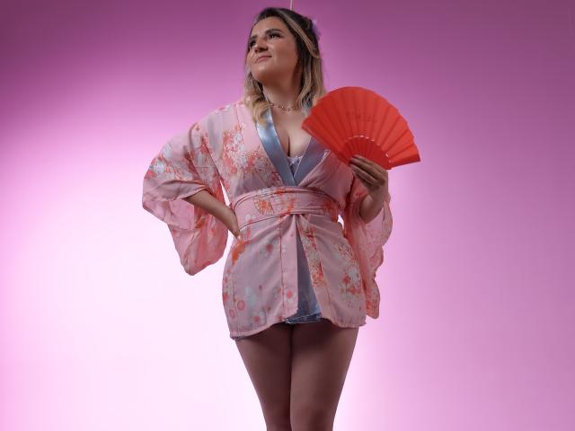 LiliEvanss in a pink floral kimono, holding a red fan, with her hand on her hip, showing her tits, against a pink gradient background.