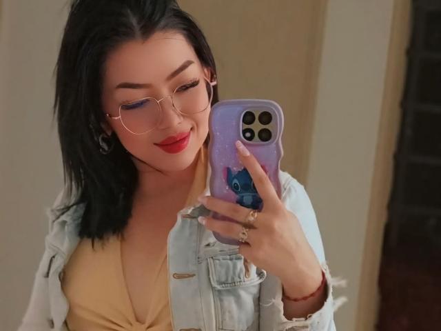 MeganKristi with black hair, red lipstick, glasses, and a denim jacket, taking a mirror selfie with a purple phone, showing off her yellow top and ring.