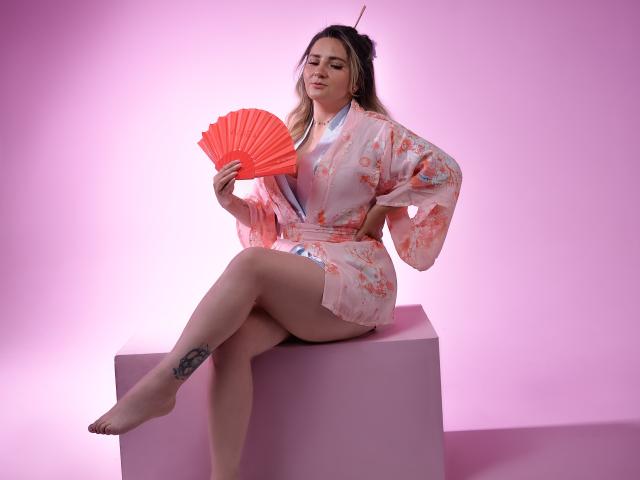 LiliEvanss in a pink floral kimono, holding an orange fan, sitting on a pink cube, with her leg up, showing a tattoo on her right thigh, pink background.