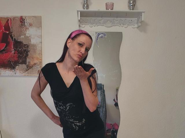 BettyO - uma mulher atraente com longos cabelos pretos, uma faixa rosa na cabeça, um vestido preto com decote em V, segurando o cabelo, em pé em frente a um espelho ondulado. Há uma prateleira branca com copos de vidro acima. A parte inferior do corpo não está visível..