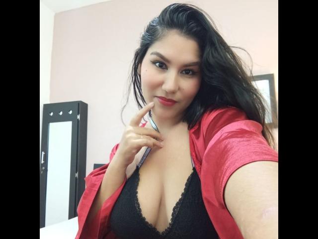 MariaRowe with long black hair, red silky robe, and black lace bra, showing off her tits with a finger to her lips, smiling slightly. She's holding a selfie.